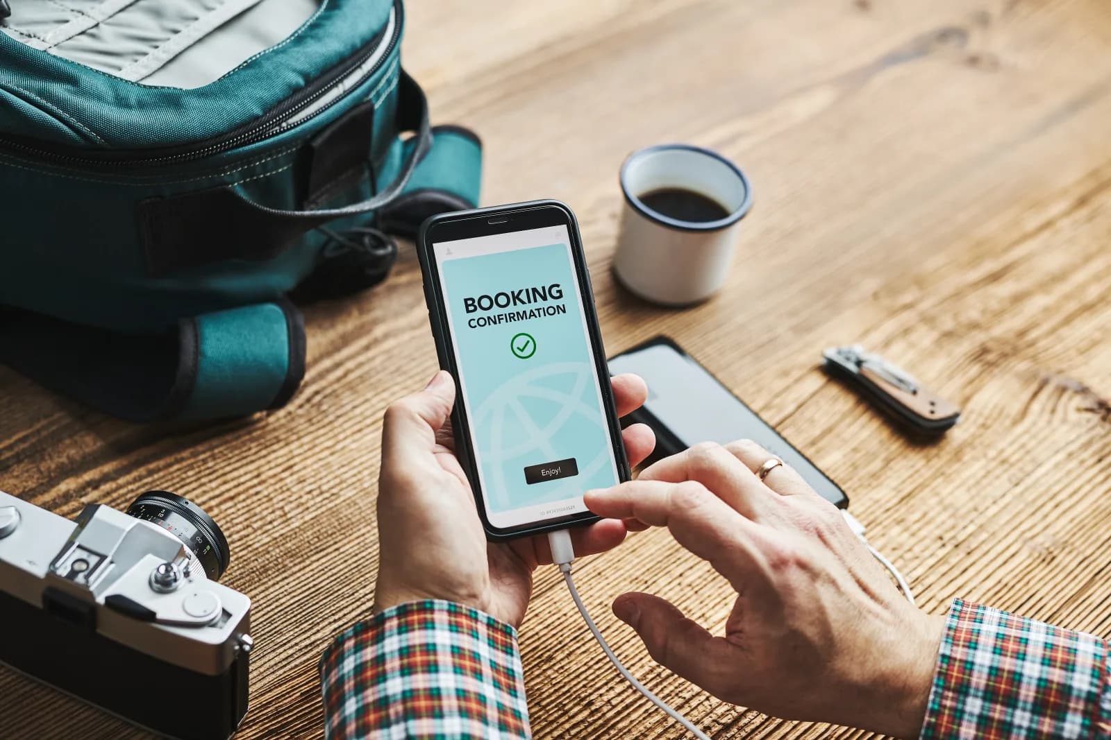 Hands holding a phone with a booking confirmation, beside a vintage camera and travel backpack on a wood table